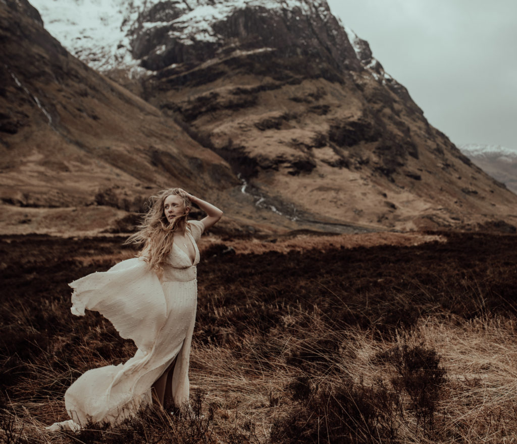 photo of the bride standing in front of the snowy mountain with wind blowing her hair and the sleeves of her dress
