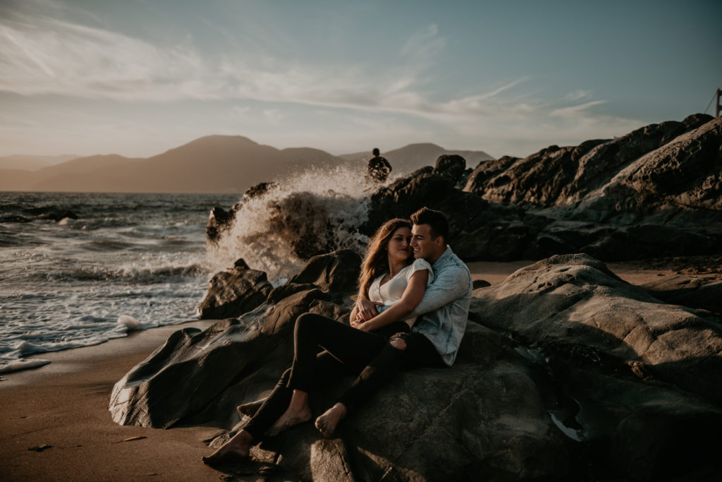 Photo of the couple sitting on the rocks at the beach with the water splashing on the rocks. 