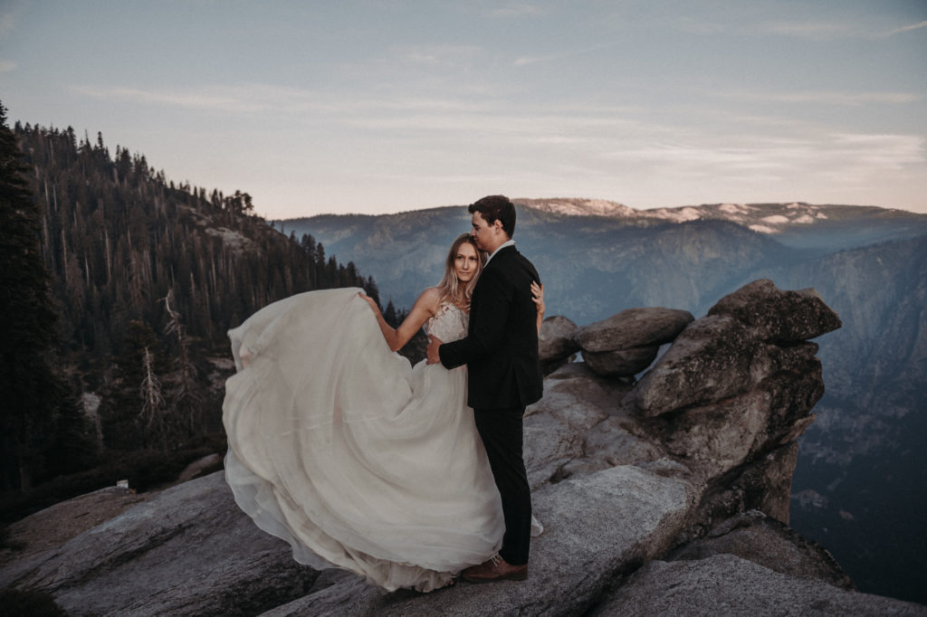 Bride holding her dress and groom hugging the bride taken on a cliff at Glacier Point at Yosemite National Park