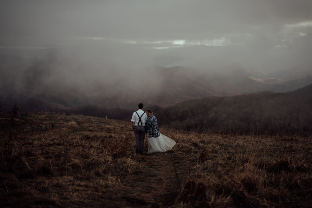 bride and groom walking away from the camera on top of a cloudy mountain during their elopement in north carolina