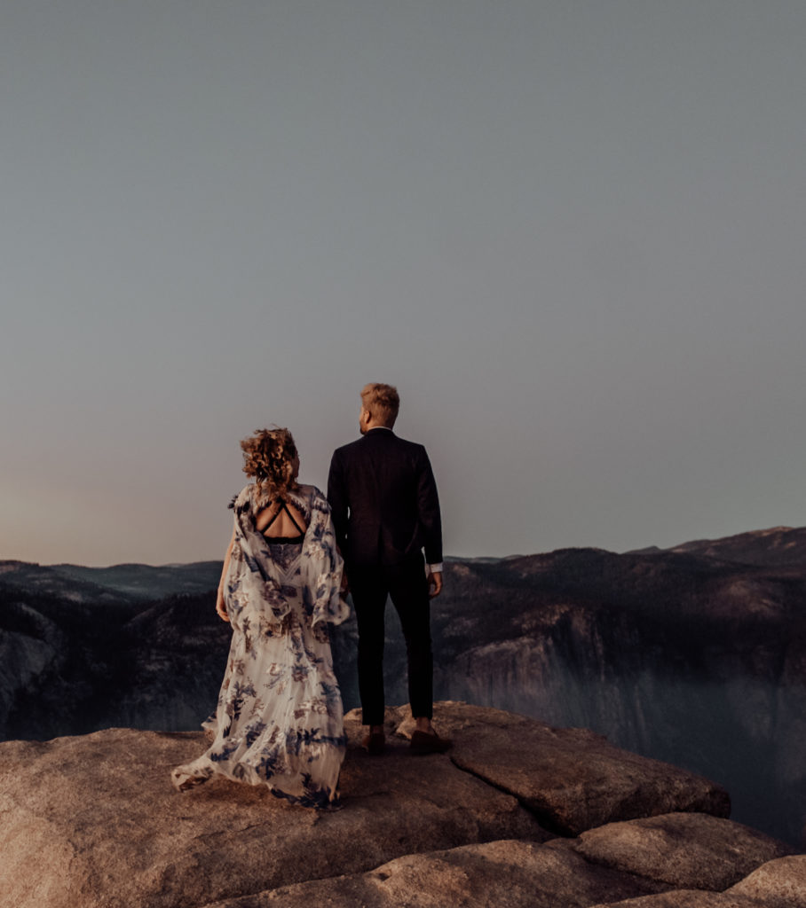 bride and groom standing on the cliff, holding hands and looking out at the vallery taken at taft point in yosemite national park