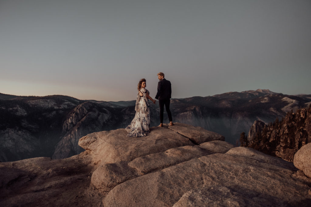 bride and groom holding hands, bride looking back & groom looking at the bride on top of a mountain at Yosemite in California