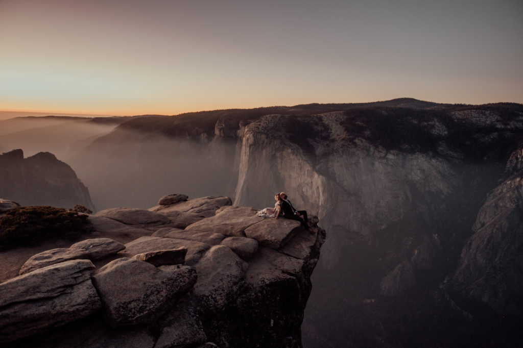 bride and groom sitting back to back at the edge of the cliff at the famous iconic yosemite spot. 
