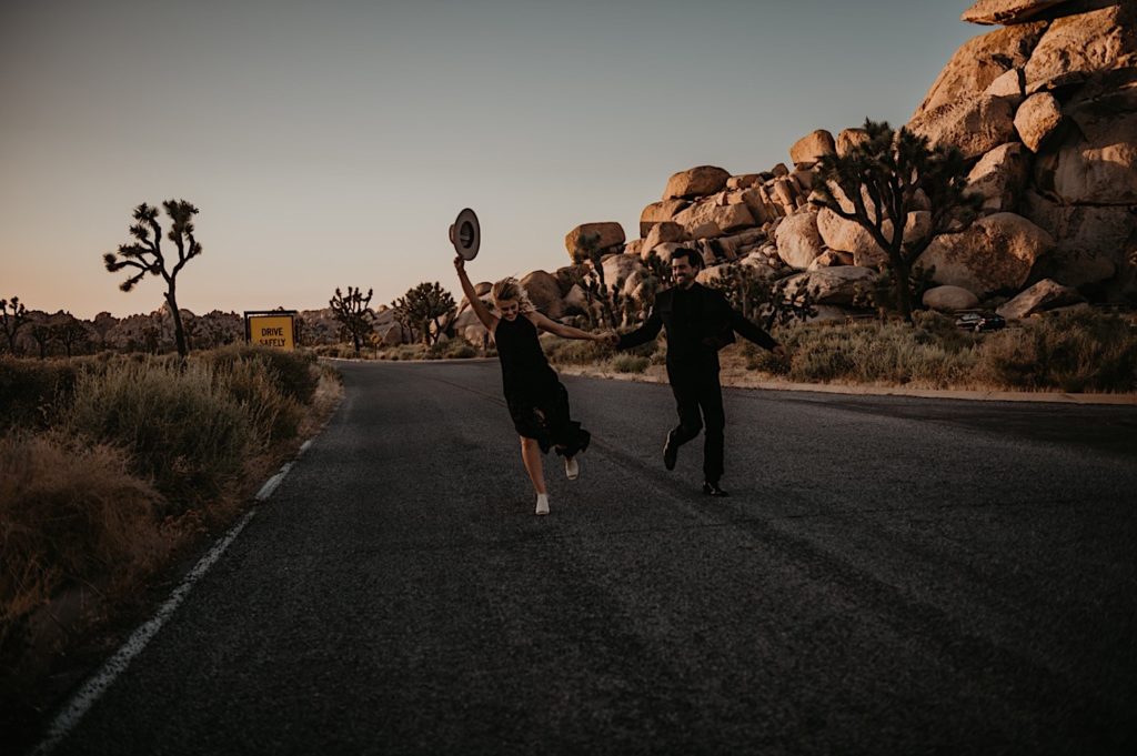 Bride and groom holding hands and running with the bride holding her hat up taken at a Joshua tree elopement