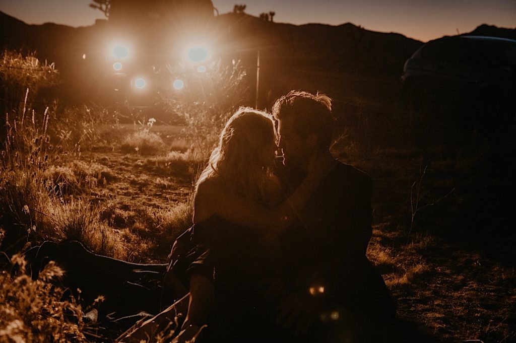 night photo of the bride and groom sitting on the ground in front of the jeep light taken at a Joshua tree elopement