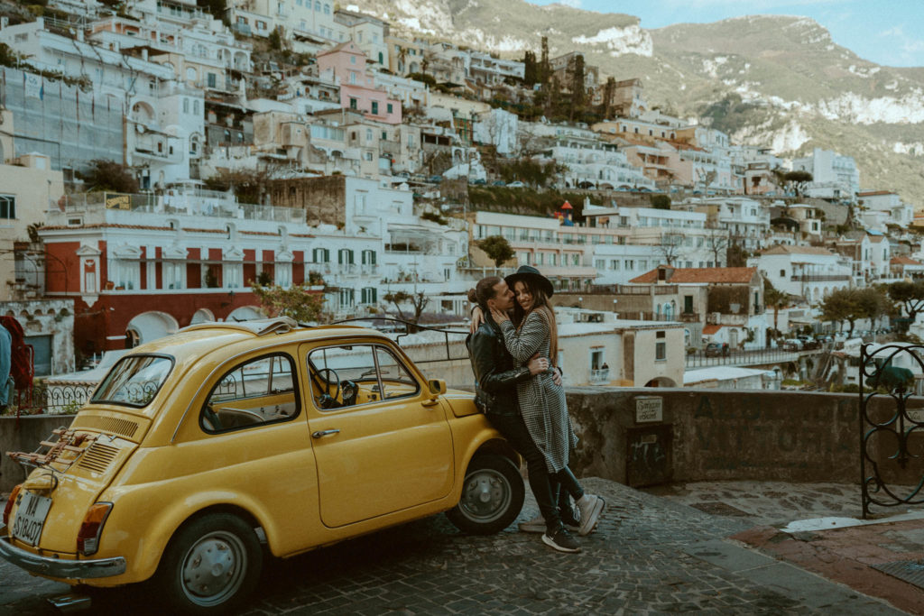 Photo of the couple hugging and sitting on the front of a vintage yellow car taken in Positano