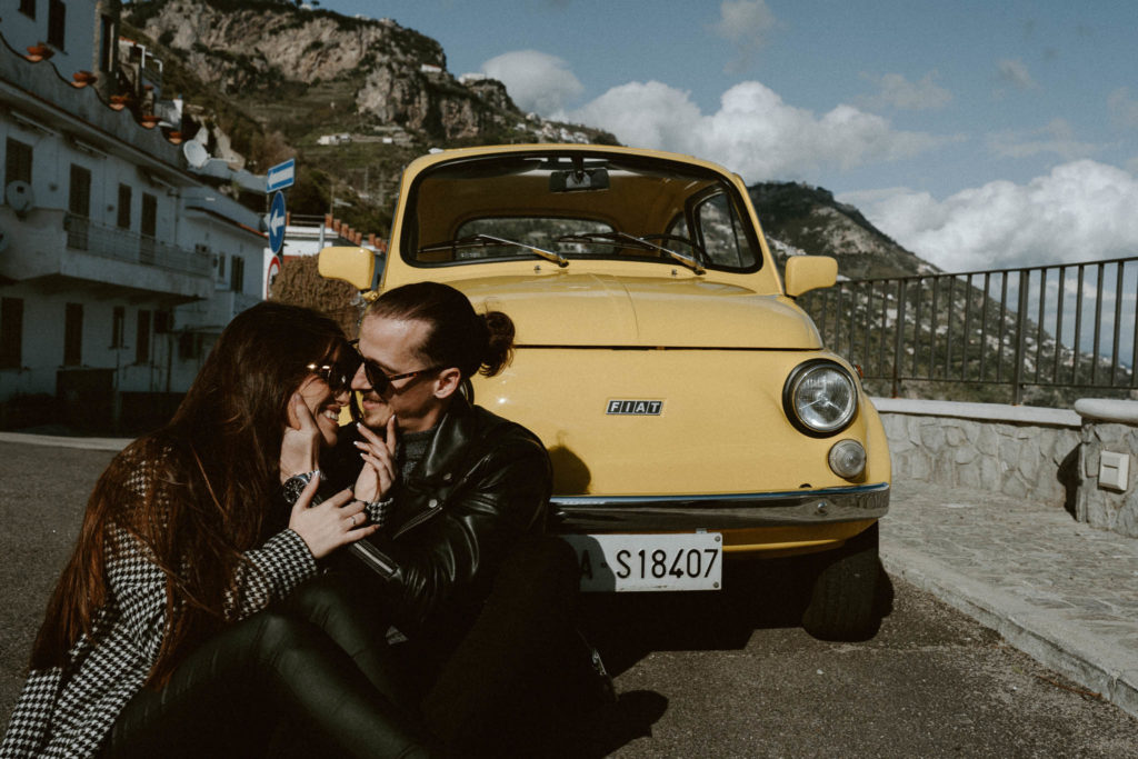 Couple sitting on the ground and looking at each other in front of a vintage yellow car in Italy