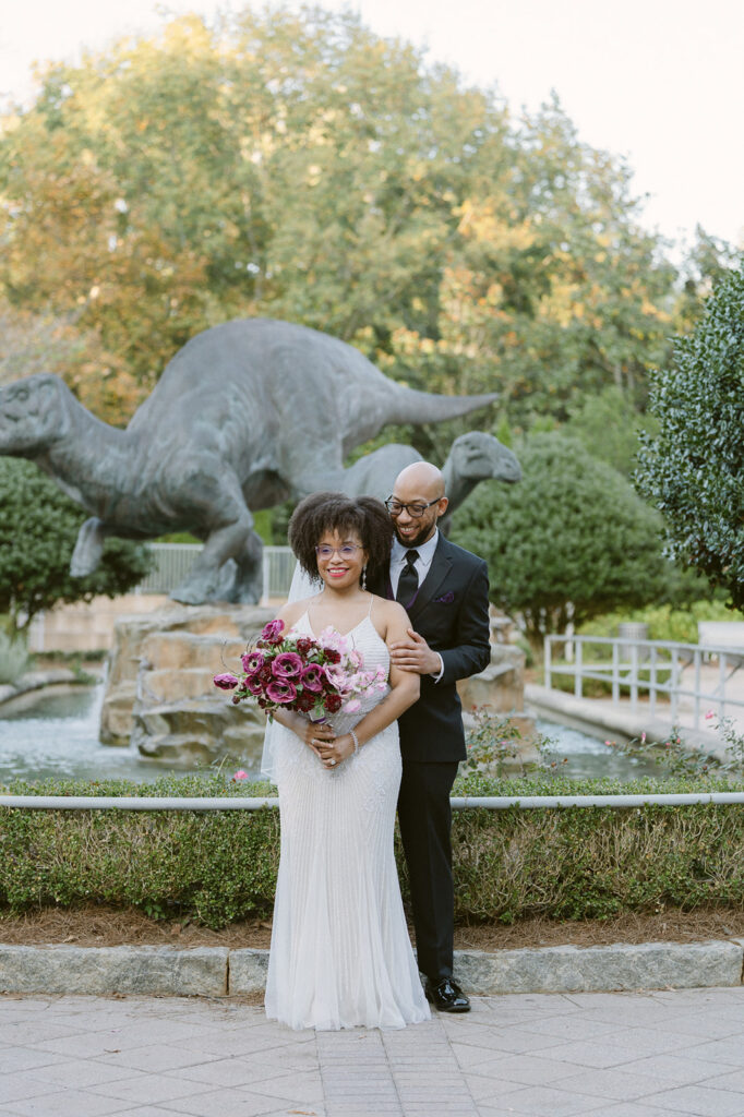 Bride and groom posing in front of dinosaur sculptures for their Fernbank Museum wedding photos in Atlanta, Georgia. 