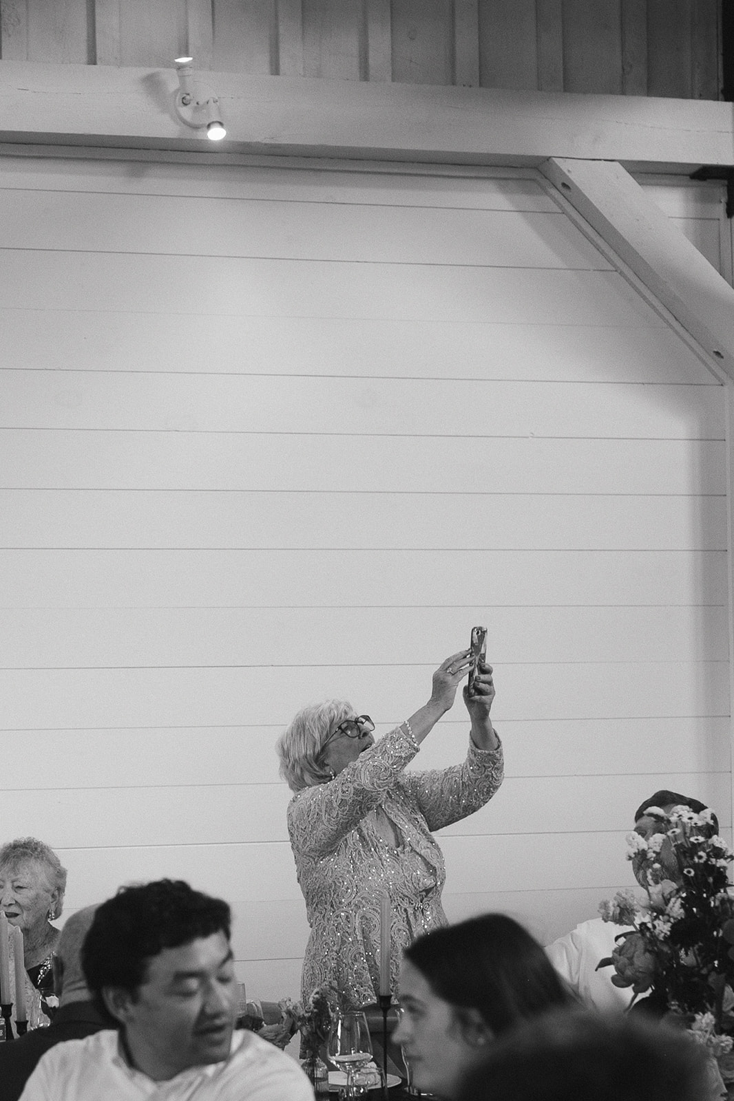Black and white photo of a wedding guest taking a photo of the bride and groom as they enter the wedding reception 