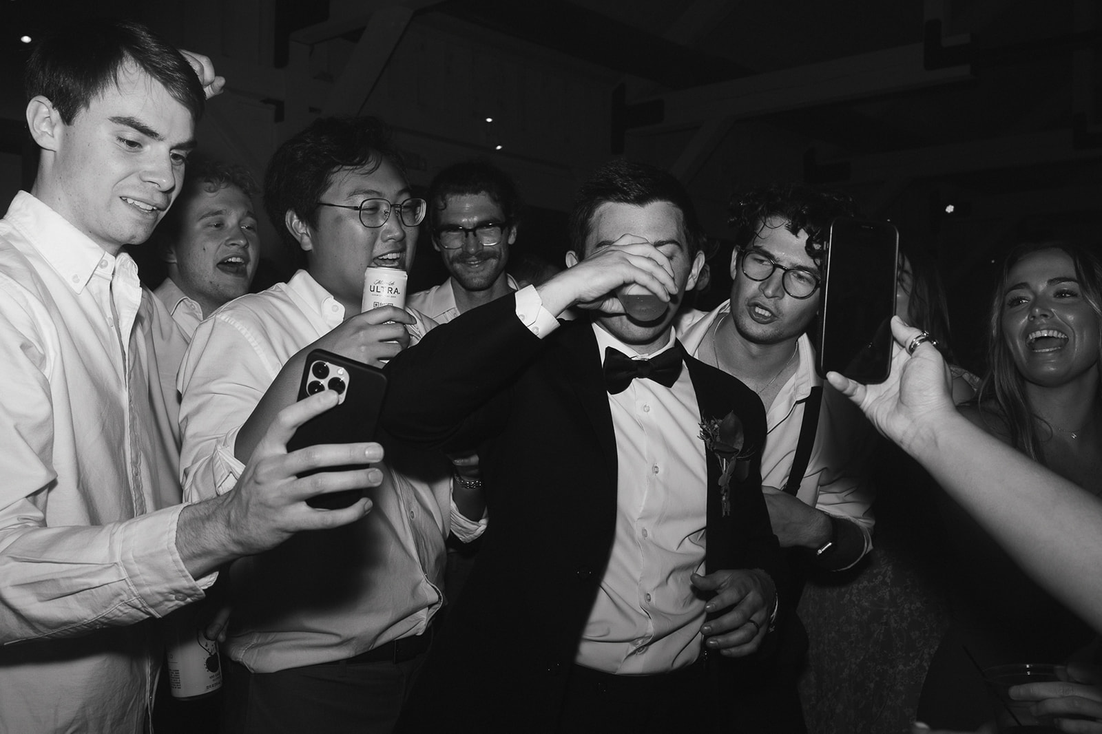 Black and white photo of a groom taking a drink during his wedding reception surrounded by groomsmen.